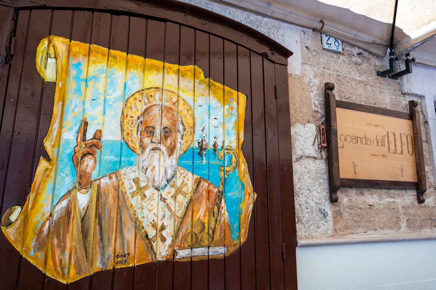 Portone in legno dipinto con santo benedicente e velieri, accanto all'insegna Locanda dell'Elfo su muro in pietra